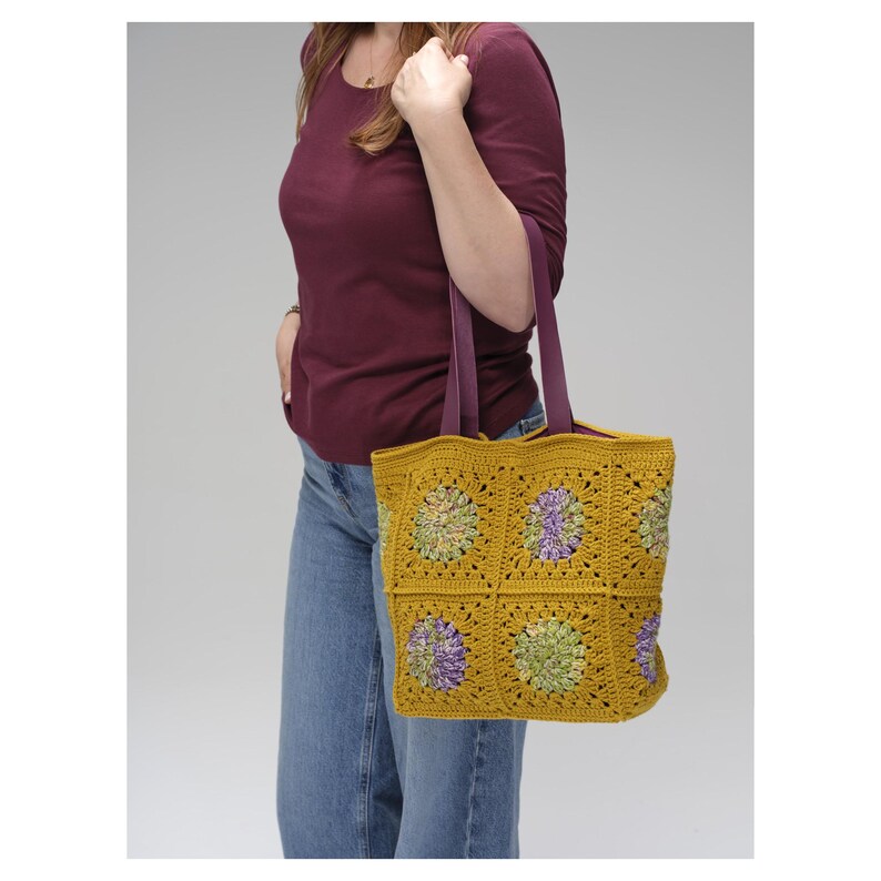 Puede incluir: Bolso tote de crochet amarillo mostaza con detalles florales morados y verdes. El bolso presenta un dise&ntilde;o de patr&oacute;n cuadrado y correas moradas. El bolso es llevado por una persona que viste una camisa burdeos y vaqueros azules.