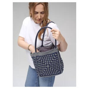 May include: A navy blue and grey crocheted tote bag with a geometric pattern. The bag has dark blue handles and a grey trim. The interior of the bag is a solid grey fabric. The bag is held by a person wearing a white t-shirt and blue jeans.