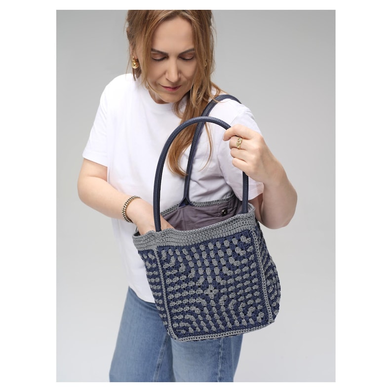 May include: A navy blue and grey crocheted tote bag with a geometric pattern. The bag has dark blue handles and a grey trim. The interior of the bag is a solid grey fabric. The bag is held by a person wearing a white t-shirt and blue jeans.