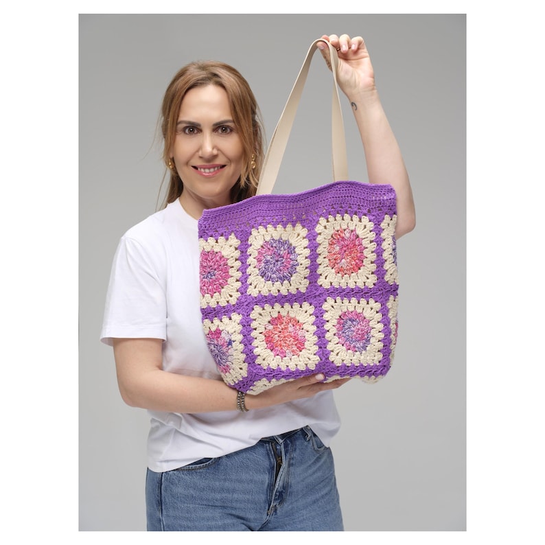 May include: A handmade crochet tote bag with a vibrant purple border and cream-colored squares, each featuring a floral design in pink and peach. The bag has cream-colored handles and is held up by a person.