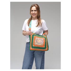K&ouml;nnte beinhalten: Eine farbenfrohe H&auml;keltasche mit Granny-Square-Design in Gr&uuml;n, Orange und Rosa. Die Tasche hat einen t&uuml;rkisfarbenen Riemen mit Holzknopfdetails. Die Tasche wird von einer Person getragen, die ein wei&szlig;es T-Shirt und blaue Jeans tr&auml;gt.