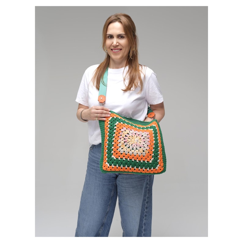 K&ouml;nnte beinhalten: Eine farbenfrohe H&auml;keltasche mit Granny-Square-Design in Gr&uuml;n, Orange und Rosa. Die Tasche hat einen t&uuml;rkisfarbenen Riemen mit Holzknopfdetails. Die Tasche wird von einer Person getragen, die ein wei&szlig;es T-Shirt und blaue Jeans tr&auml;gt.