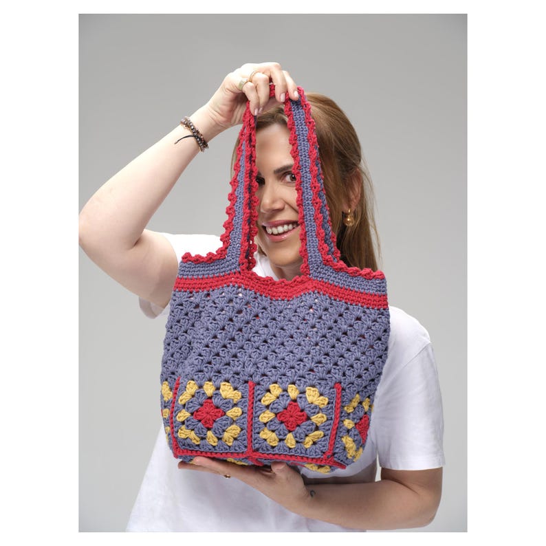 May include: A handmade crochet tote bag in a blue-gray color with red and yellow accents. The bag features a detailed stitch pattern and decorative squares on the front. The handles and top trim are red, adding a pop of color to the design.