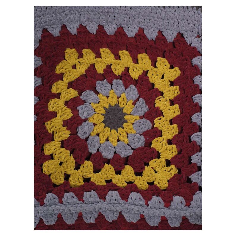 May include: A square crochet textile featuring a central dark gray circle surrounded by concentric layers of light gray, yellow, and deep red yarn. The pattern consists of repeating clusters of stitches forming a geometric granny square design.