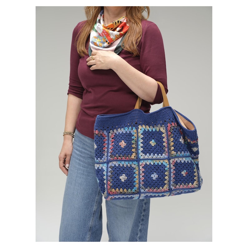 Puede incluir: Un bolso tote de ganchillo azul con un dise&ntilde;o de cuadrados de la abuela. El bolso presenta una base azul oscuro con cuadrados coloridos y asas de cuero marr&oacute;n claro. El bolso est&aacute; abierto en la parte superior y parece espacioso.