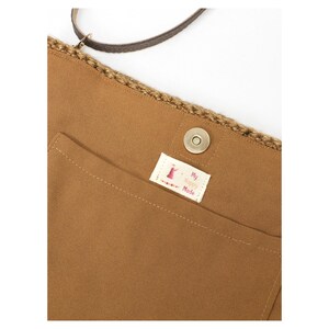 May include: A tan fabric bag featuring a crocheted top edge, a dark brown strap, and a magnetic snap closure. A small white rectangular label is sewn onto the front pocket, displaying the text My Happily Made in red and black lettering.
