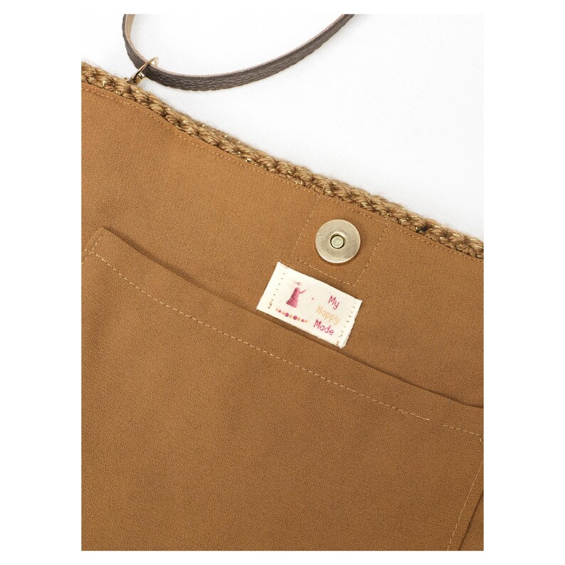 May include: A tan fabric bag featuring a crocheted top edge, a dark brown strap, and a magnetic snap closure. A small white rectangular label is sewn onto the front pocket, displaying the text My Happily Made in red and black lettering.