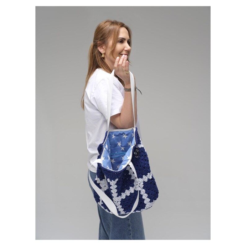 May include: A navy blue and white crochet tote bag with a white strap. The bag's interior is lined with blue fabric and white stars. The bag is carried over the shoulder.
