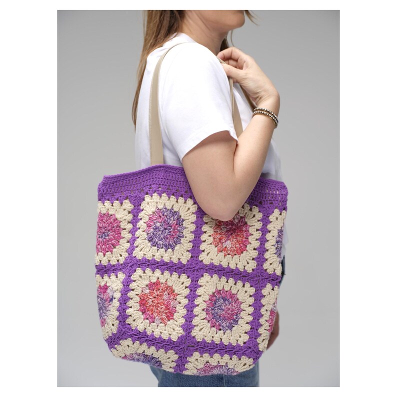May include: A handmade crochet tote bag with a purple border and cream-colored squares. Each square features a floral design with pink, red, and purple accents. The bag has beige straps and is carried over the shoulder.