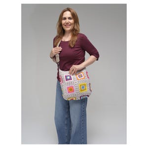 May include: A beige crochet shoulder bag with colourful square patterns. The squares feature various colours, including pink, yellow, blue, and purple. The bag has a long strap and is held by a person wearing a burgundy top and blue jeans.