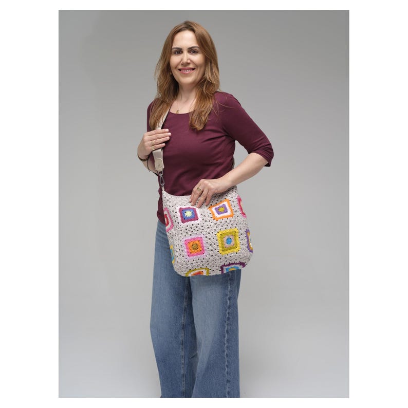 May include: A beige crochet shoulder bag with colourful square patterns. The squares feature various colours, including pink, yellow, blue, and purple. The bag has a long strap and is held by a person wearing a burgundy top and blue jeans.