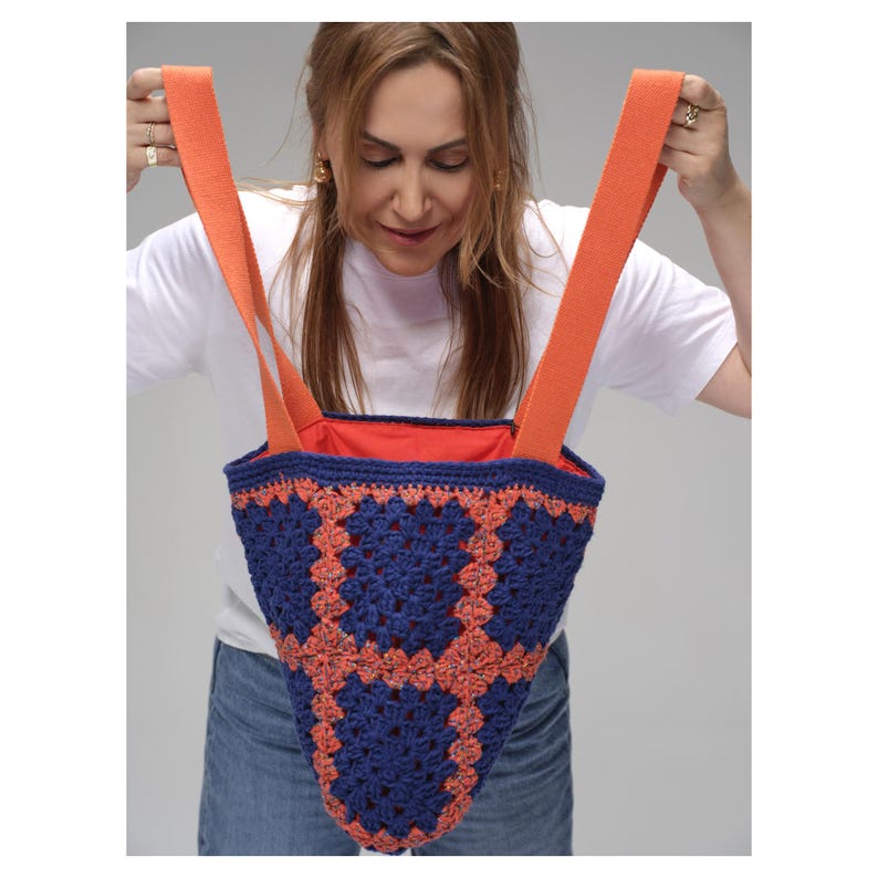 May include: A handmade crochet tote bag with a blue and orange geometric pattern. The bag has orange straps and a bright orange interior. The bag is held up by a person.