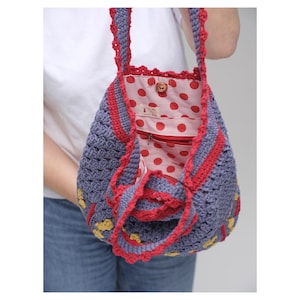 May include: A handmade crochet bag in shades of blue, red, and yellow. The bag features a red polka dot lining and a red zipper. The straps and edges are trimmed with red crochet. The bag is held by a person wearing a white shirt and blue jeans.