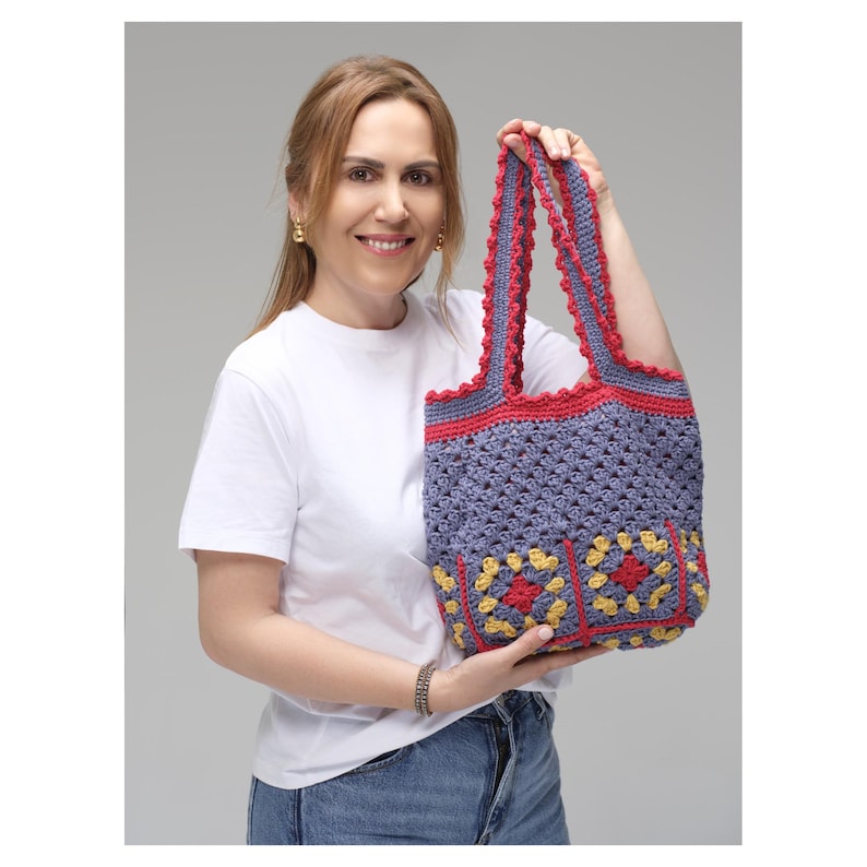 May include: A handmade crochet tote bag in shades of blue, red, and yellow. The bag features a granny square design on the front and a scalloped edge. The bag is held up by a person.