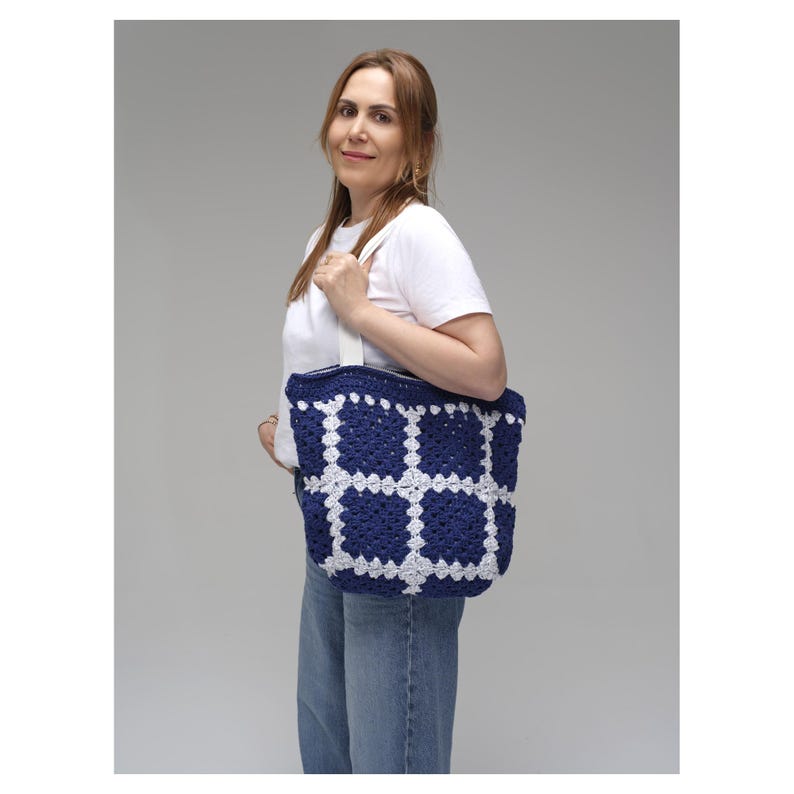 May include: A navy blue and white crochet tote bag with a grid pattern. The bag has a rectangular shape and white straps. The bag is held by a person wearing a white t-shirt and blue jeans.