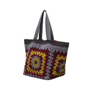 May include: A crochet tote bag featuring a granny square pattern in shades of burgundy, mustard yellow, and light gray. The bag has two gray fabric handles and a dark gray crocheted top border, with a visible red interior lining at the opening.
