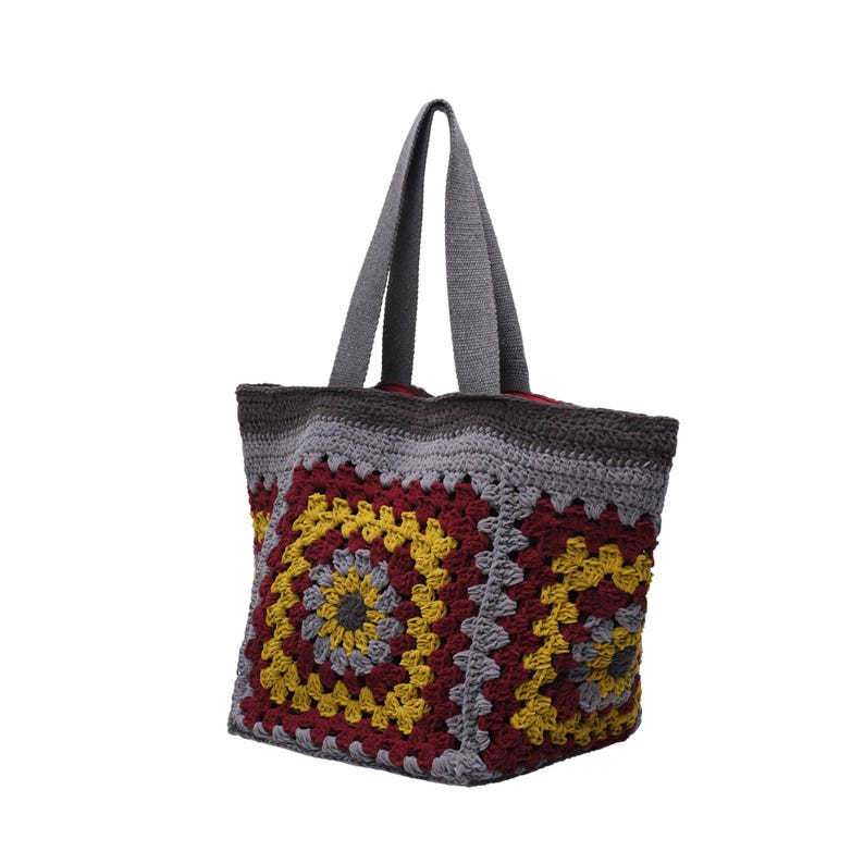 May include: A crochet tote bag featuring a granny square pattern in shades of burgundy, mustard yellow, and light gray. The bag has two gray fabric handles and a dark gray crocheted top border, with a visible red interior lining at the opening.