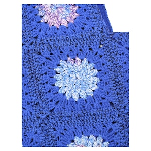 May include: Close-up of a blue crocheted item with floral patterns. The flowers are in shades of blue, purple, and white. The item has a textured appearance, with a visible stitch pattern. The overall design is intricate and handmade.