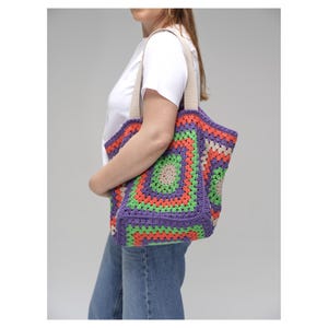Puede incluir: Un bolso tote de ganchillo colorido con un patr&oacute;n geom&eacute;trico. El bolso presenta un dise&ntilde;o cuadrado con tonos de morado, naranja, verde y beige. Tiene correas beige y se lleva al hombro.