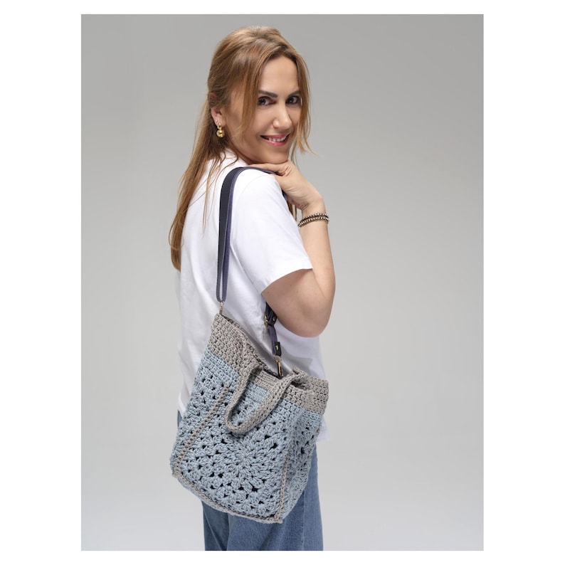 May include: A crocheted tote bag in shades of blue and gray, with a dark blue strap. The bag has a textured, open weave design and a gray top border. The bag is being carried over the shoulder.