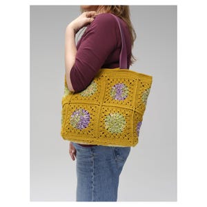Puede incluir: Bolso tote de crochet amarillo mostaza con detalles florales morados y verdes. El bolso presenta un dise&ntilde;o de patr&oacute;n cuadrado y una correa morada. El bolso tote es un art&iacute;culo hecho a mano.