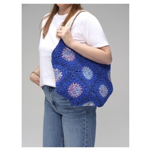 May include: A blue crochet tote bag with a floral pattern in shades of blue, purple, and white. The bag has a cream-colored strap and is held by a person. The bag is a handmade item.