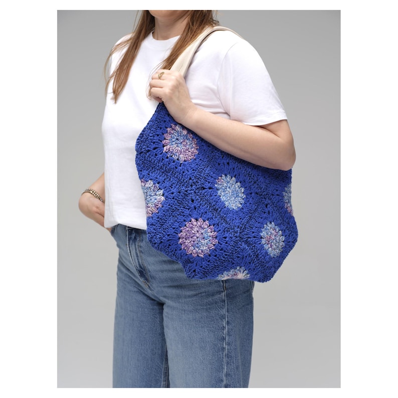 May include: A blue crochet tote bag with a floral pattern in shades of blue, purple, and white. The bag has a cream-colored strap and is held by a person. The bag is a handmade item.