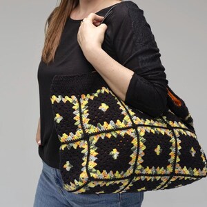 Puede incluir: Un bolso tote de crochet negro con un colorido estampado floral en amarillo, verde y blanco. El bolso lo sostiene una persona que lleva una camisa negra de manga larga y vaqueros azules. El bolso tiene forma rectangular y un asa resistente.