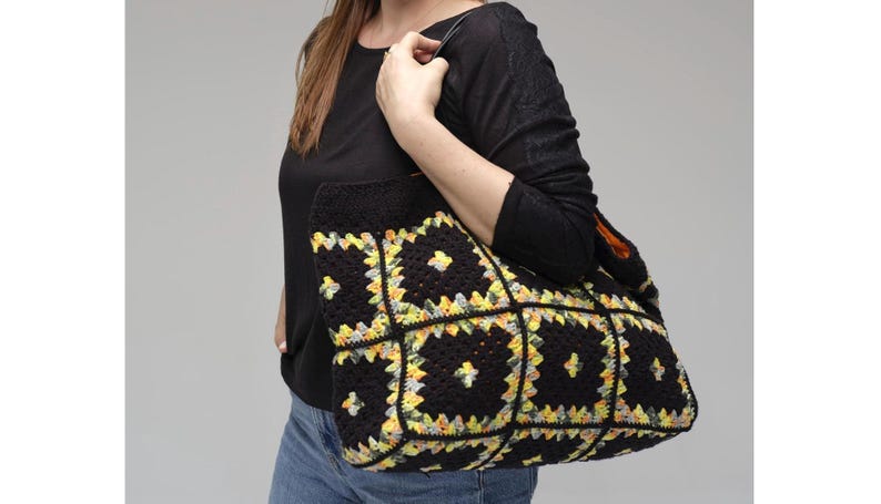 Puede incluir: Un bolso tote de crochet negro con un colorido estampado floral en amarillo, verde y blanco. El bolso lo sostiene una persona que lleva una camisa negra de manga larga y vaqueros azules. El bolso tiene forma rectangular y un asa resistente.