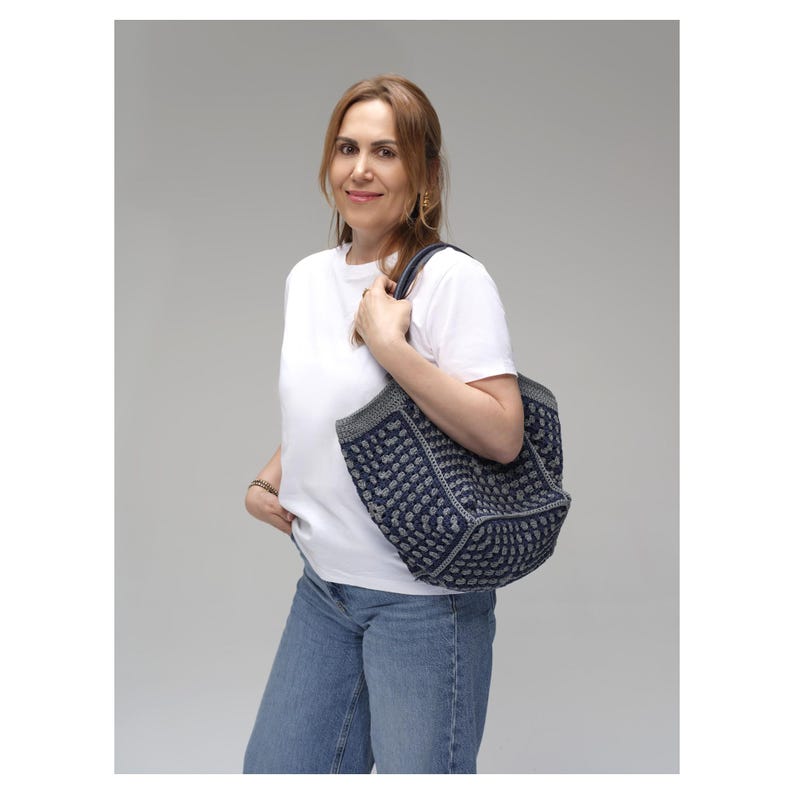 May include: A navy blue tote bag with a white geometric pattern is held by a person. The bag has a grey trim and handles. The person is wearing a white t-shirt and blue jeans.