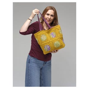 Puede incluir: Un bolso tote de crochet amarillo hecho a mano con asas moradas. El bolso presenta un estampado floral en tonos verdes y morados. El bolso de mano est&aacute; sostenido por una persona.