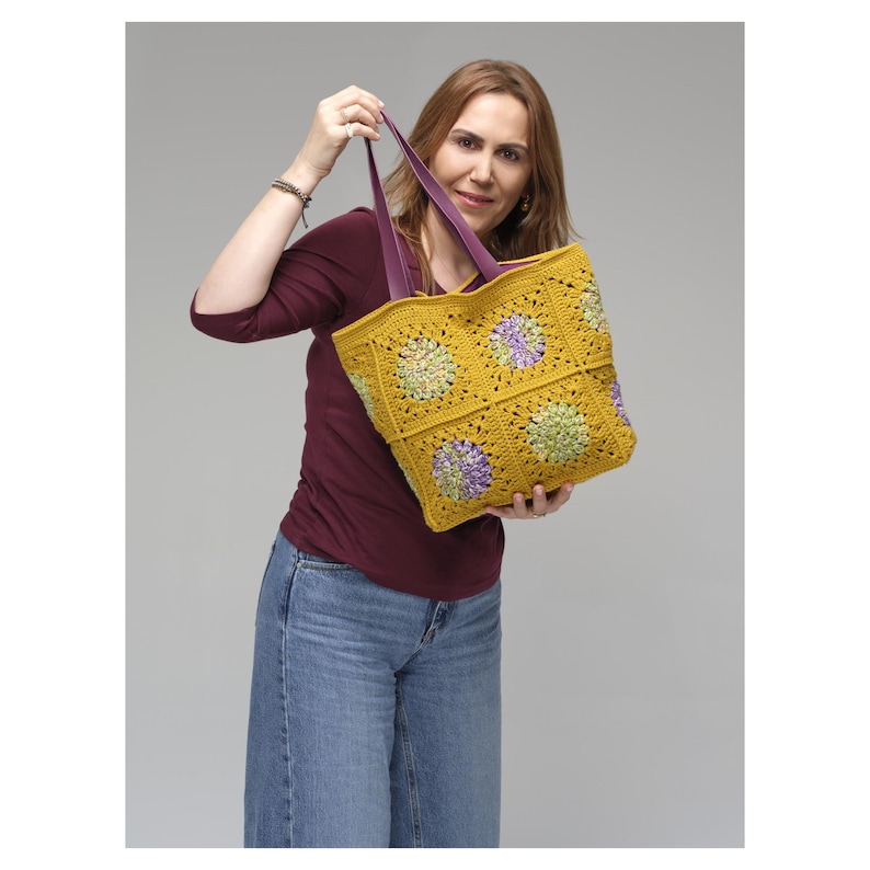 Puede incluir: Un bolso tote de crochet amarillo hecho a mano con asas moradas. El bolso presenta un estampado floral en tonos verdes y morados. El bolso de mano est&aacute; sostenido por una persona.