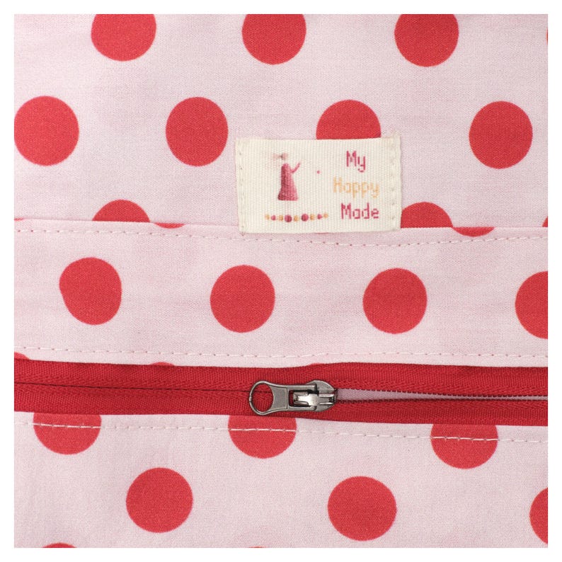 May include: Close-up of a pink fabric with large red polka dots. A red zipper runs horizontally across the bottom. A small label with the text "My Happy Made" and a graphic of a person is sewn on the fabric.