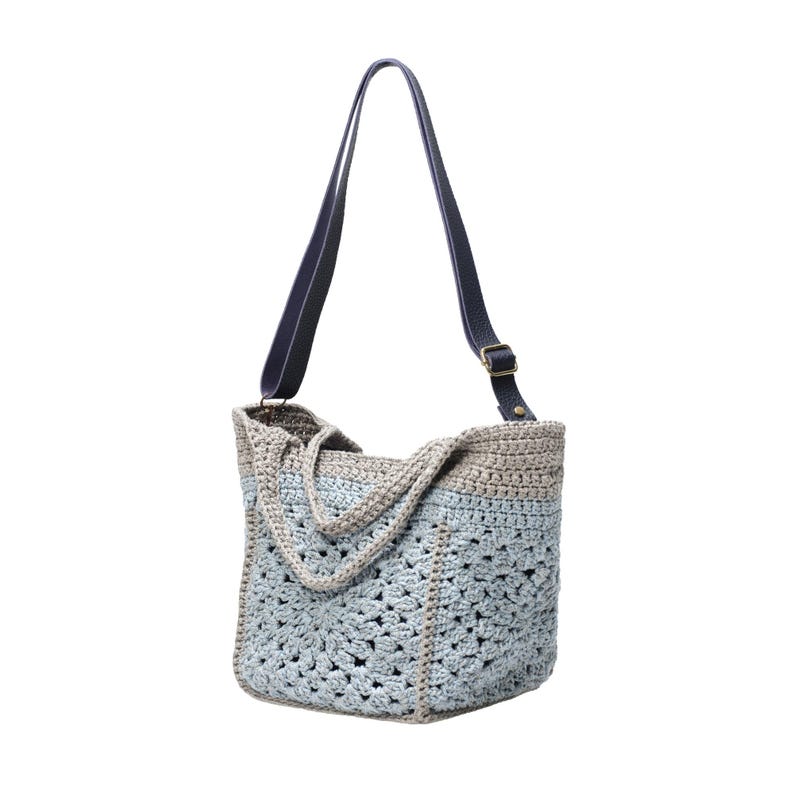 May include: A handmade crochet tote bag in shades of blue and gray, featuring a geometric openwork design. The bag has a dark blue leather strap with a brass buckle. The tote is designed for carrying items.