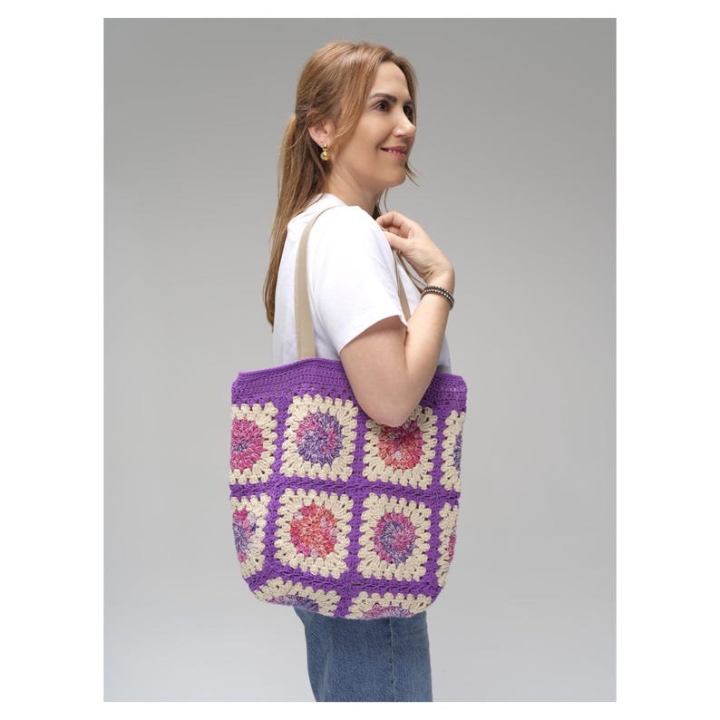May include: A handmade crochet tote bag with a purple border and a patchwork design. The squares feature a mix of pink, cream, and purple colors. The bag has beige straps and is carried over the shoulder.