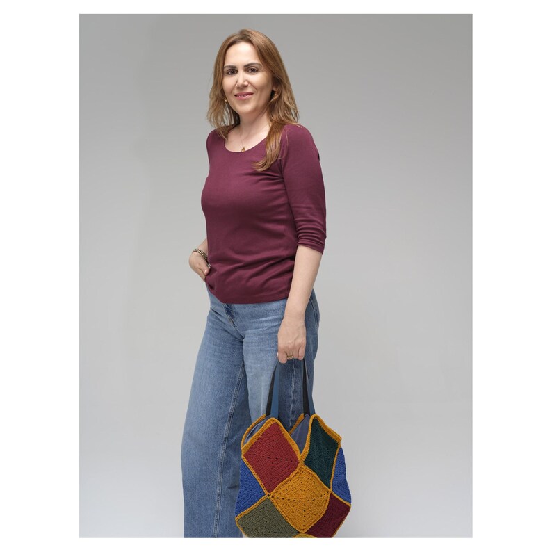 Puede incluir: Una mujer con una camisa burdeos de manga larga y vaqueros azules, sosteniendo un bolso tote de ganchillo patchwork colorido con asas negras. El bolso presenta cuadrados rojos, amarillos, azules y verdes.