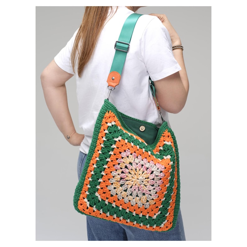 K&ouml;nnte beinhalten: Bunte H&auml;keltasche mit Schulterriemen, mit einem Granny-Square-Muster in Gr&uuml;n, Orange und Wei&szlig;. Die Tasche hat ein gr&uuml;nes Innenfutter und einen verstellbaren, t&uuml;rkisfarbenen Riemen mit orangefarbenen Lederdetails. Die Tasche wird &uuml;ber der Schulter getragen.