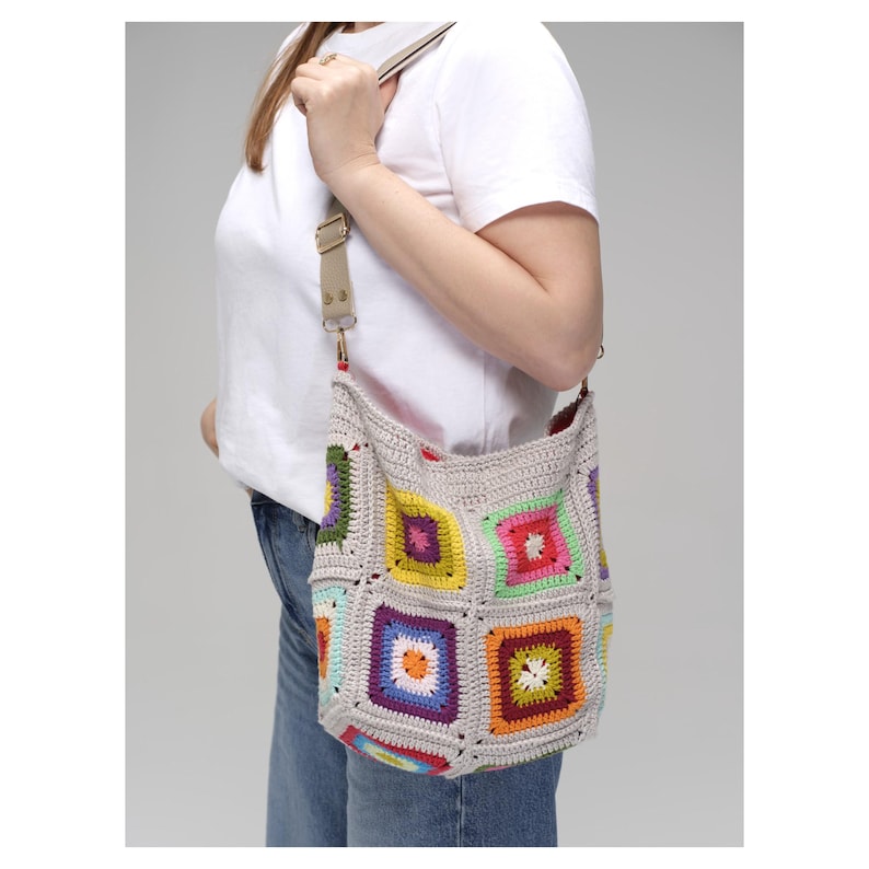K&ouml;nnte beinhalten: Eine geh&auml;kelte Umh&auml;ngetasche mit einem farbenfrohen Patchwork-Design. Die Tasche hat Quadrate in verschiedenen Farben, darunter Gr&uuml;n, Gelb, Orange und Blau, vor einem neutralen grauen Hintergrund. Sie hat einen beigefarbenen, verstellbaren Riemen.