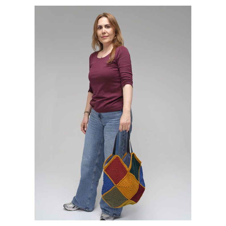 Puede incluir: Una camisa burdeos de manga larga y vaqueros azules. Se sostiene un bolso tote patchwork con asa negra. El bolso tote presenta cuadrados rojos, amarillos, azules y verdes.