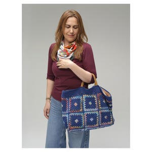 Puede incluir: Una mujer sostiene un gran bolso tote azul tejido a mano. El bolso presenta un dise&ntilde;o de patchwork con cuadrados coloridos. Tiene asas de cuero marr&oacute;n y un interior espacioso.