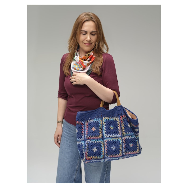 Puede incluir: Una mujer sostiene un gran bolso tote azul tejido a mano. El bolso presenta un dise&ntilde;o de patchwork con cuadrados coloridos. Tiene asas de cuero marr&oacute;n y un interior espacioso.