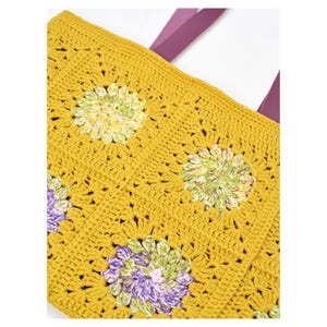 Puede incluir: Bolso tote de crochet amarillo mostaza con un estampado floral en tonos verdes, morados y blancos. El bolso presenta un dise&ntilde;o de cuadrados de la abuela y correas moradas. Este bolso hecho a mano es perfecto para llevar la compra o art&iacute;culos esenciales.