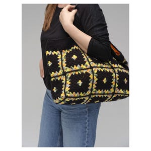 Puede incluir: Bolso tote de crochet negro con un colorido estampado floral en amarillo, verde y gris. El bolso lo lleva una persona con una camisa negra de manga larga y vaqueros azules. El bolso tiene un bolsillo grande.