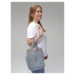 May include: A handmade crochet tote bag in shades of light blue and gray. The bag features a textured, open weave design with sturdy handles. The bag is carried over the shoulder, showcasing its casual style and craftsmanship.