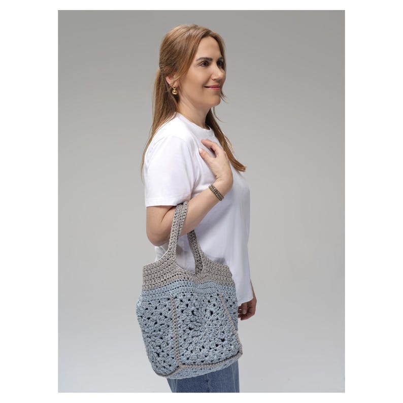 May include: A handmade crochet tote bag in shades of light blue and gray. The bag features a textured, open weave design with sturdy handles. The bag is carried over the shoulder, showcasing its casual style and craftsmanship.