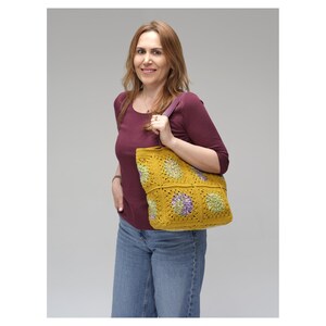 Puede incluir: Un bolso tote de crochet amarillo mostaza con una correa morada. El bolso presenta un patr&oacute;n cuadrado con dise&ntilde;os florales verdes y morados. La mujer de la imagen lleva una camisa burdeos y vaqueros azules.