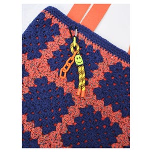 May include: Close-up of a handmade crochet tote bag with a granny square pattern in navy blue and coral. The bag has orange straps and a yellow and orange smiley face keychain attached with a black carabiner.