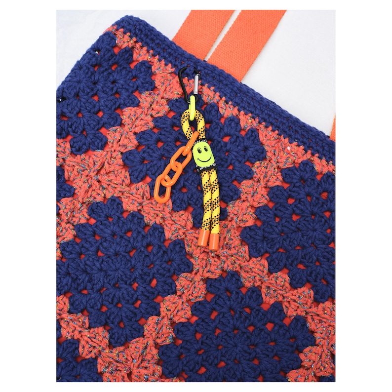 May include: Close-up of a handmade crochet tote bag with a granny square pattern in navy blue and coral. The bag has orange straps and a yellow and orange smiley face keychain attached with a black carabiner.