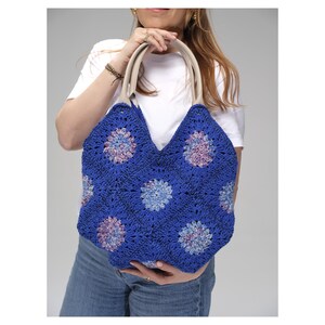 May include: A blue crochet tote bag with a floral pattern. The bag has cream-colored handles and a V-shaped top. The floral pattern features shades of blue, purple, and pink. The bag is held by a person wearing a white t-shirt and blue jeans.