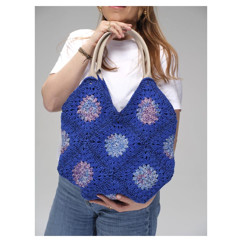 May include: A blue crochet tote bag with a floral pattern. The bag has cream-colored handles and a V-shaped top. The floral pattern features shades of blue, purple, and pink. The bag is held by a person wearing a white t-shirt and blue jeans.